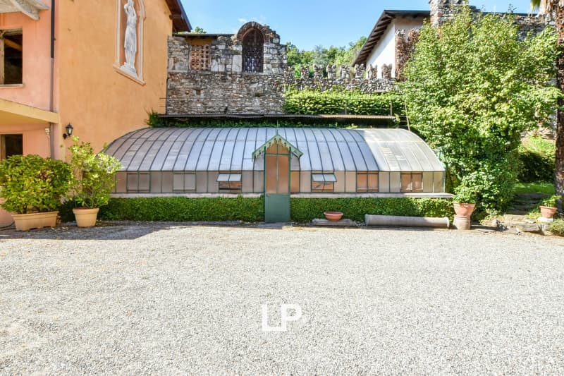 A stone building with a large greenhouse structure attached, surrounded by lush greenery and a gravel courtyard.