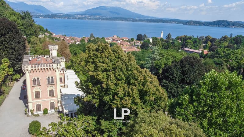 Aerial view of a lakeside town surrounded by mountains and lush greenery. The image shows a historic castle-like building, a church tower, and a scenic lake with boats. The town is nestled among trees and surrounded by the mountainous landscape.