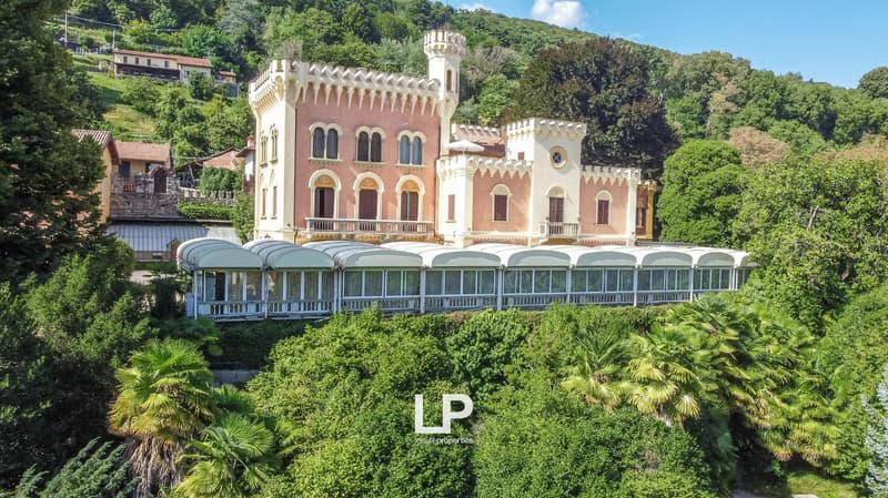 A large, ornate castle-like building with a pink and white facade, towers, and arched windows. The building is surrounded by lush greenery and palm trees, with a covered terrace or balcony area in the front.