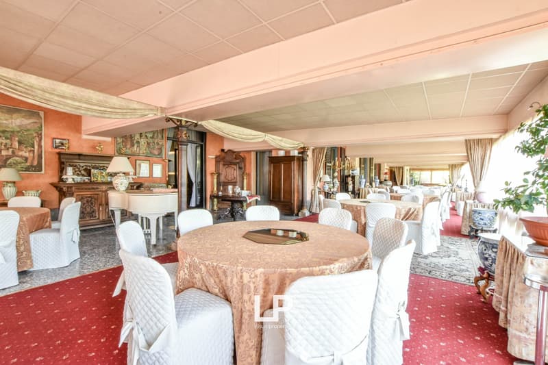 Large dining room with a round table covered in a patterned tablecloth, surrounded by white upholstered chairs. The room has a high ceiling with decorative curtains, and the walls are decorated with artwork and antique furniture pieces.