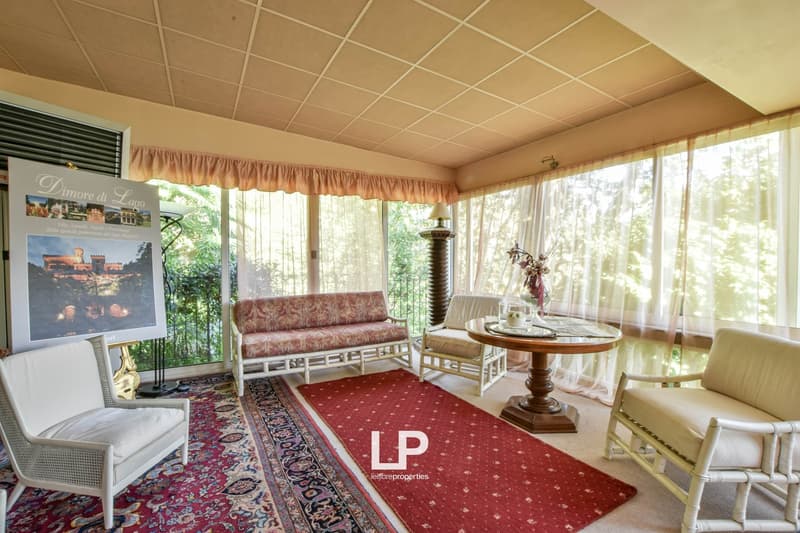 Cozy living room with large windows overlooking a balcony or terrace, featuring a floral patterned sofa, a round wooden table, and a decorative rug. The room has a warm, inviting atmosphere with curtains and a tiled ceiling.