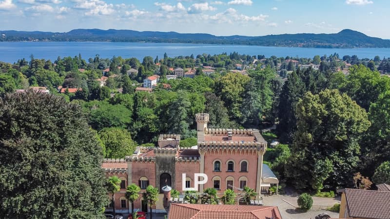 The image shows a scenic view of a lakeside town surrounded by lush greenery and mountains. The town features a mix of residential and commercial buildings, including a large castle-like structure with red-tiled roofs and towers. The lake in the backgroun