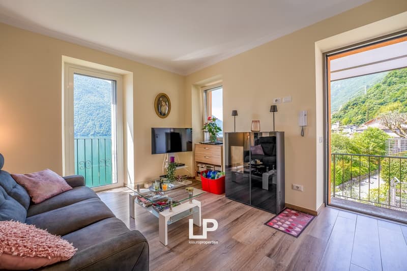 Spacious living room with hardwood floors, beige walls, large windows with a balcony overlooking the scenic mountain view, a gray sofa, a glass coffee table, and a TV mounted on the wall. The room also features a dresser and some decor items.