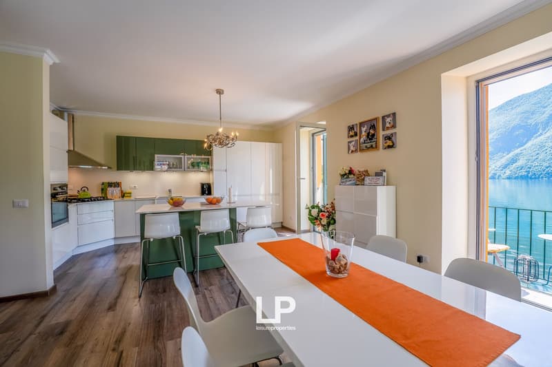 Open-concept kitchen and dining area with large windows offering a scenic view of the lake. The kitchen features white cabinets, green accents, and modern appliances. The dining table has an orange table runner and is surrounded by white chairs. The room