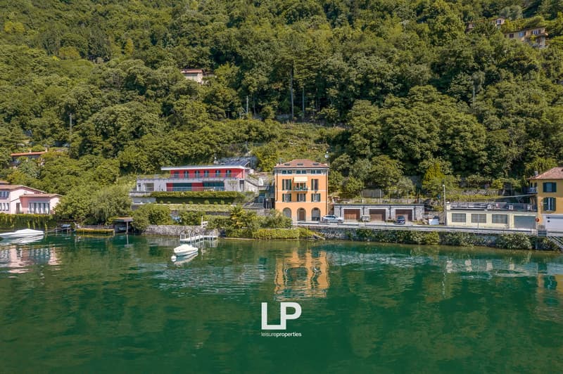 Colorful buildings with balconies overlooking a calm, turquoise lake surrounded by lush, green trees and vegetation. Several boats and docks are visible on the lake's surface.