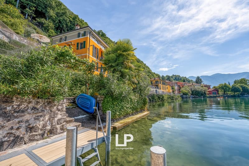 Colorful multi-story building with balconies, surrounded by lush greenery and palm trees, situated along a lake with a wooden dock and boats in the water
