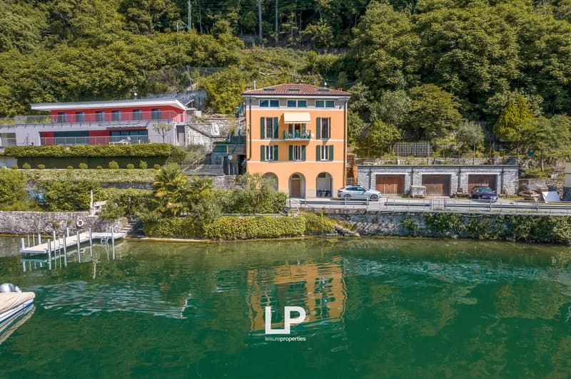 Large house with yellow walls, multiple windows, balcony with chairs, parked car in front, garage next to the house, surrounded by greenery and mountains, dock and boat in the water.