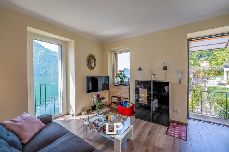 Spacious living room with large windows offering a scenic view of the lake and mountains, hardwood flooring, beige walls, sofa, coffee table, TV stand, and balcony access