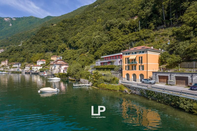 Picturesque lakeside town with colorful buildings, boats, and lush green mountains in the background. The image shows a scenic waterfront area with a road running along the lake and several multi-story residential or commercial buildings with balconies an