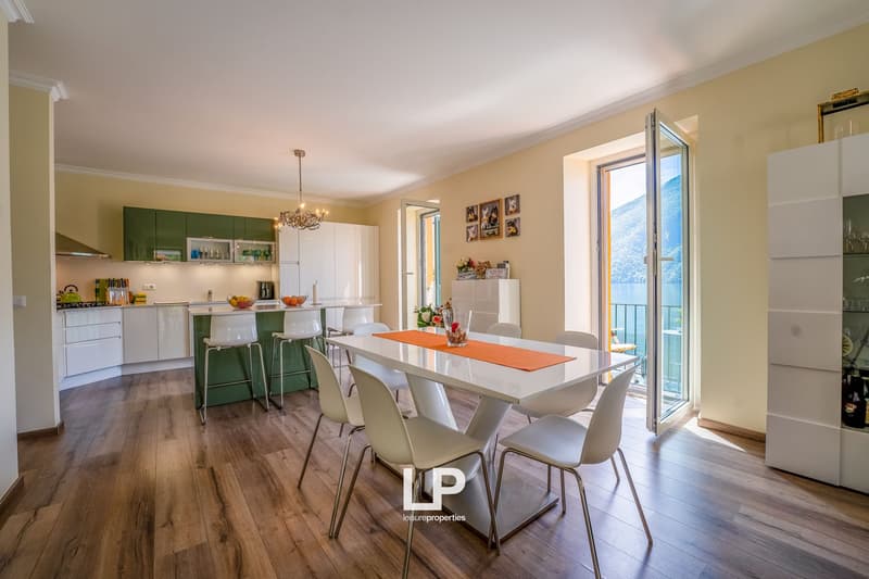 Hardwood floors, cream-colored walls, modern chandelier, white dining table with chairs, green kitchen with stools, glass doors to balcony, window overlooking a scenic view, white and green cabinets, bar counter, various kitchen appliances, glass display