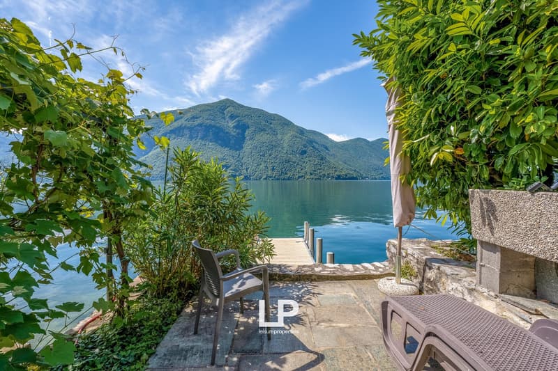 Scenic lakeside view with a wooden dock, surrounded by lush greenery and mountains in the background. There is a patio area with outdoor furniture overlooking the tranquil lake.