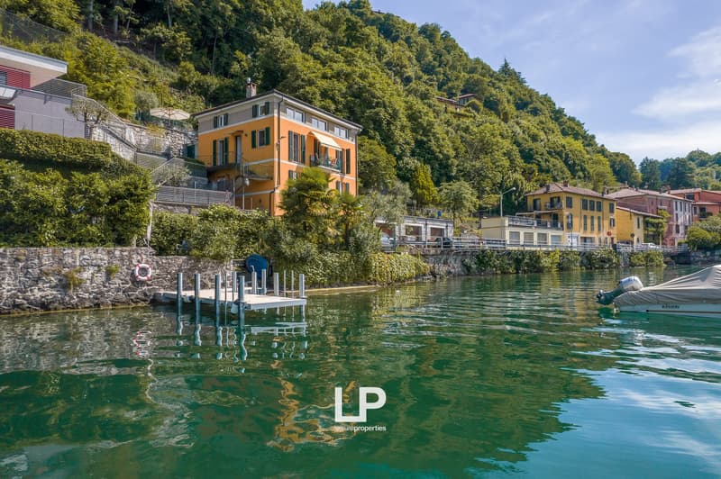Colorful multi-story houses with balconies overlooking a lake, surrounded by lush greenery and mountains. There is a dock and boats in the water.