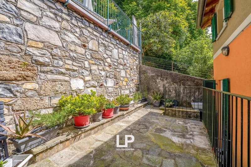 Stone wall exterior, wooden balcony railing, potted plants, green foliage surrounding the building