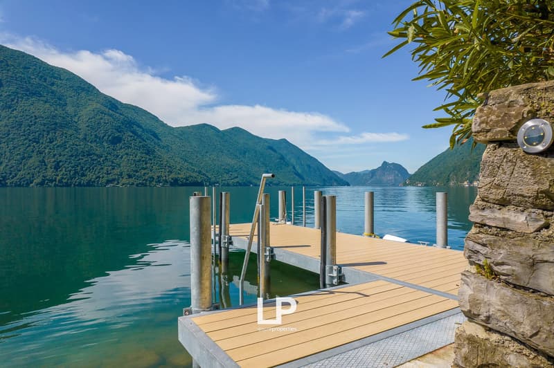 Wooden dock overlooking a scenic lake surrounded by mountains, with a clear blue sky and lush greenery in the foreground.
