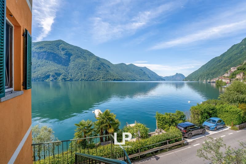 The image shows a scenic view of a lake surrounded by mountains. The lake has a calm, turquoise-blue surface reflecting the surrounding landscape. There are palm trees and other vegetation along the shore, and a road with parked cars can be seen. The buil