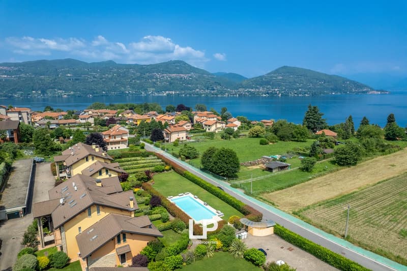 The image shows a scenic view of a residential area located on the shores of a lake, surrounded by mountains. The houses have red tile roofs and are nestled among lush greenery, including well-manicured gardens and a swimming pool. The lake and mountains