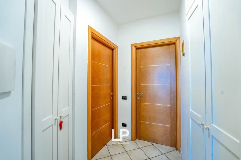 The image shows a hallway with two wooden doors, one on the left and one on the right. The walls are painted white, and the floor is covered with tile. The hallway appears to be well-lit, with natural light coming in from a window or other source.