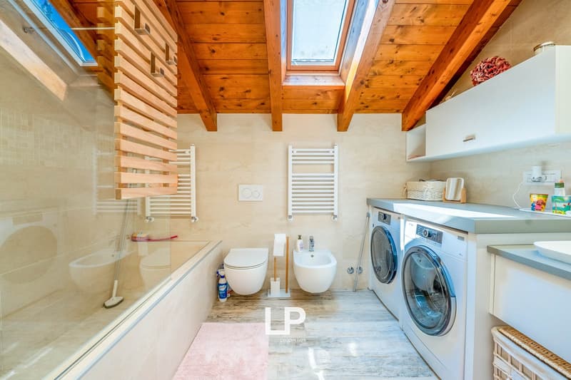 Spacious bathroom with wooden ceiling beams, tiled floor, toilet, bidet, and a washing machine and dryer set up in the room.