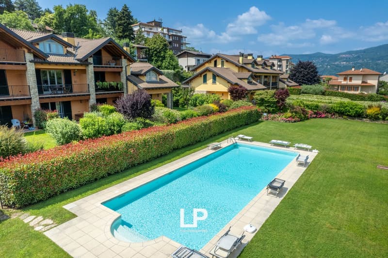 This image shows a picturesque apartment complex with multiple buildings surrounded by lush greenery and a well-manicured garden. The complex features a large outdoor swimming pool with lounge chairs around it. The buildings have a mix of architectural st