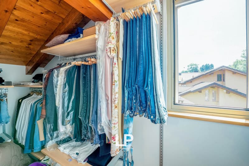 The image shows a closet or wardrobe space with various clothing items hanging on racks and shelves. The space has a wooden ceiling and large windows, providing natural light. The clothing items include dresses, shirts, and jeans in various colors and pat