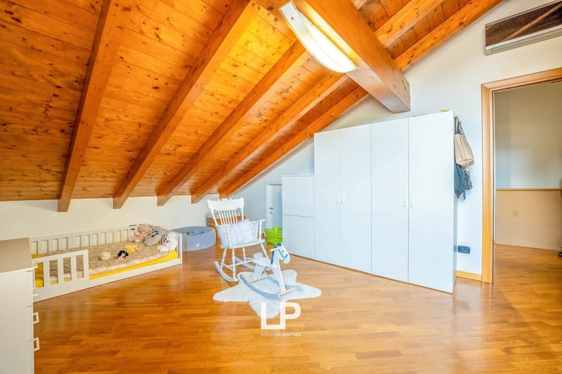 Attic bedroom with wooden beamed ceiling, hardwood floor, white cabinets, crib, rocking chair, and toys