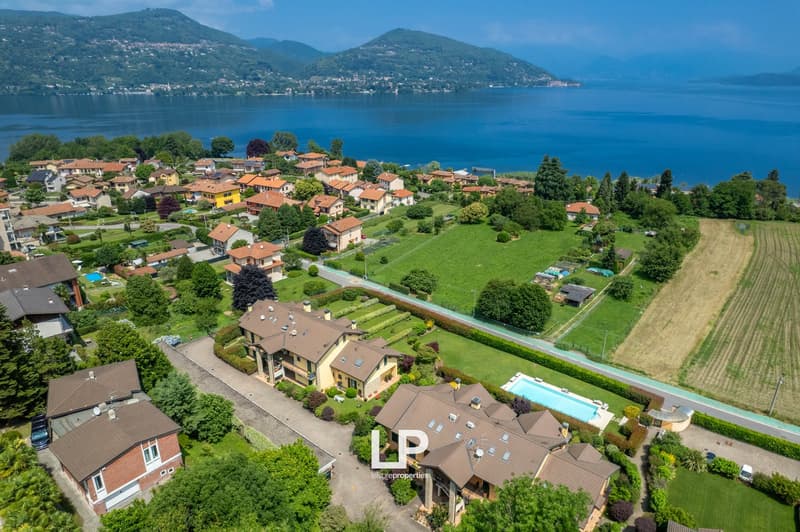 This image shows an aerial view of a picturesque lakeside town nestled among rolling hills and lush greenery. The town is composed of colorful, multi-story residential buildings with red tile roofs, surrounded by well-manicured gardens, fields, and a larg