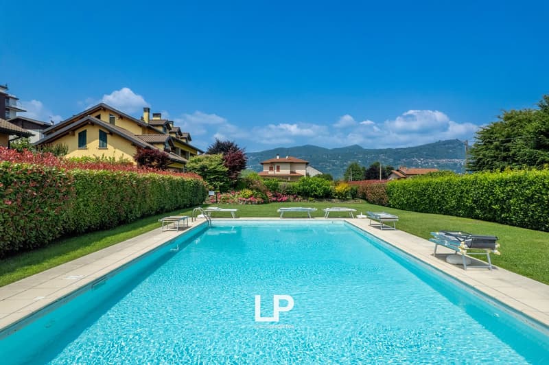 Residential complex with a large outdoor swimming pool surrounded by lush greenery, including hedges and trees. The buildings have a Mediterranean-style architecture with yellow and red-tiled roofs.