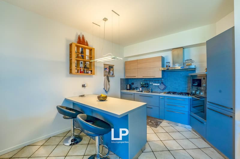 Modern kitchen with blue cabinets and drawers, white countertop, blue tile backsplash, stainless steel appliances, wooden shelving unit, and a kitchen island with bar stools.