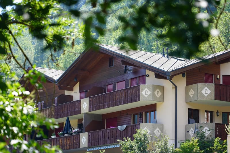 Multi-story wooden chalet-style apartment building with balconies, surrounded by lush greenery and trees