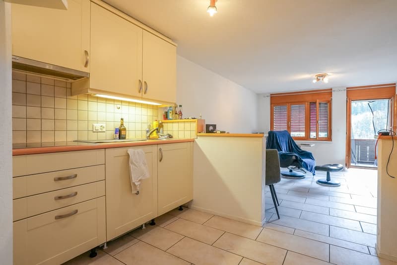 Fully equipped kitchen with beige cabinets, tile backsplash, and appliances including a washing machine and dishwasher. The kitchen opens up to a living area with a blue chair and ottoman.