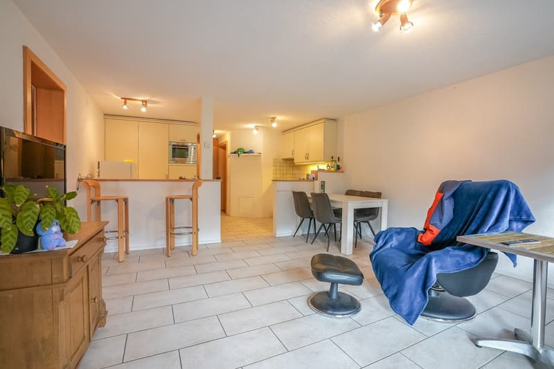 Open-concept living area with kitchen, dining table, and living room furniture. Hardwood floors, white walls, and recessed lighting. Kitchen has built-in appliances including a dishwasher.
