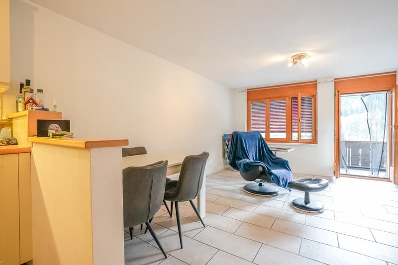 Spacious living room with tiled flooring, white walls, and large windows. The room features a wooden counter with kitchen appliances, a comfortable armchair with a blue blanket, and a dining table with chairs.