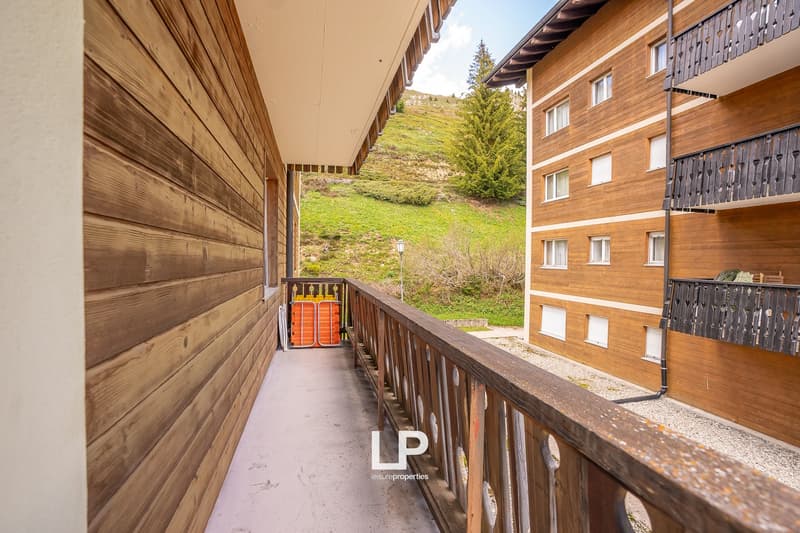 A multi-story wooden apartment building with a balcony overlooking a grassy hillside with pine trees. The building has a wooden exterior and a covered walkway along the side.
