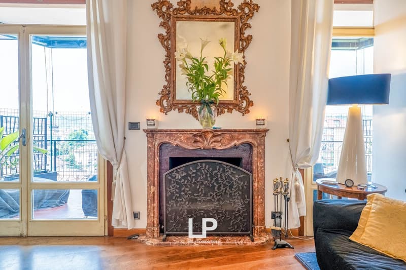 Ornate, antique-style fireplace with carved wooden frame, large mirror above, floral arrangement in vase, hardwood floors, large windows, blue sofa, and other decorative elements