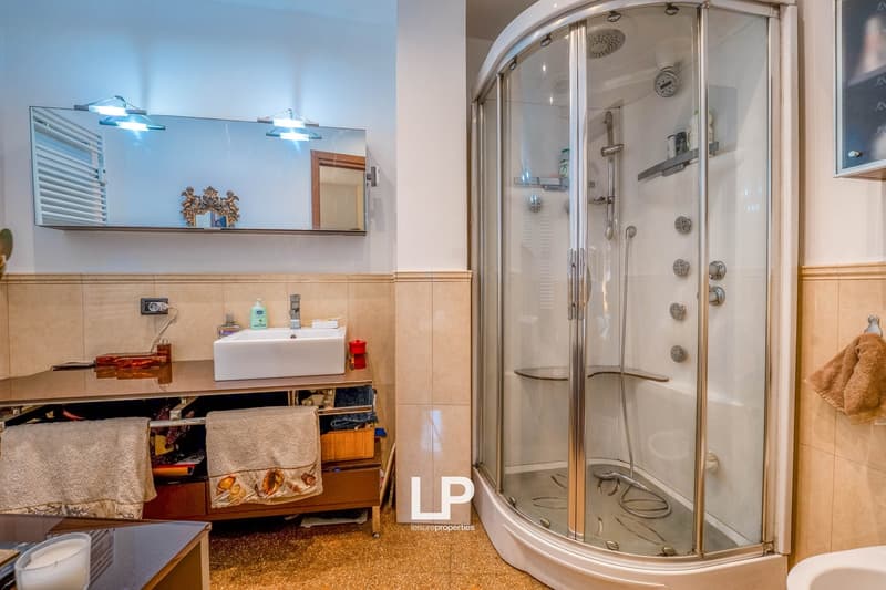 The image shows a modern bathroom with a glass-enclosed shower stall, a vanity with a sink, and storage cabinets. The bathroom has tiled walls and floors, and appears to be well-lit with recessed lighting fixtures.