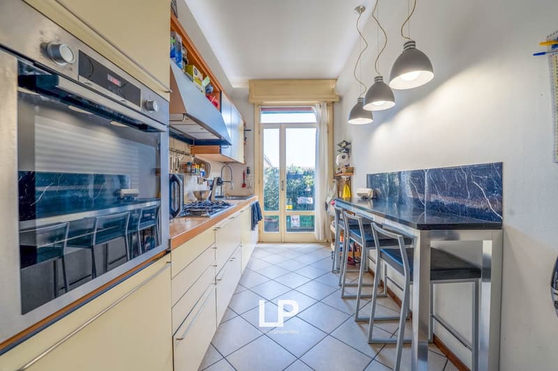 Spacious kitchen with modern appliances, including a dishwasher and washing machine. The kitchen has wooden cabinets, a tiled floor, and a large window providing natural light. There is a dining area with a table and stools adjacent to the kitchen.
