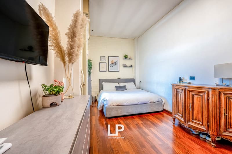 Spacious bedroom with hardwood floors, white walls, large windows, gray upholstered bed, wooden dresser, and decorative plants and artwork