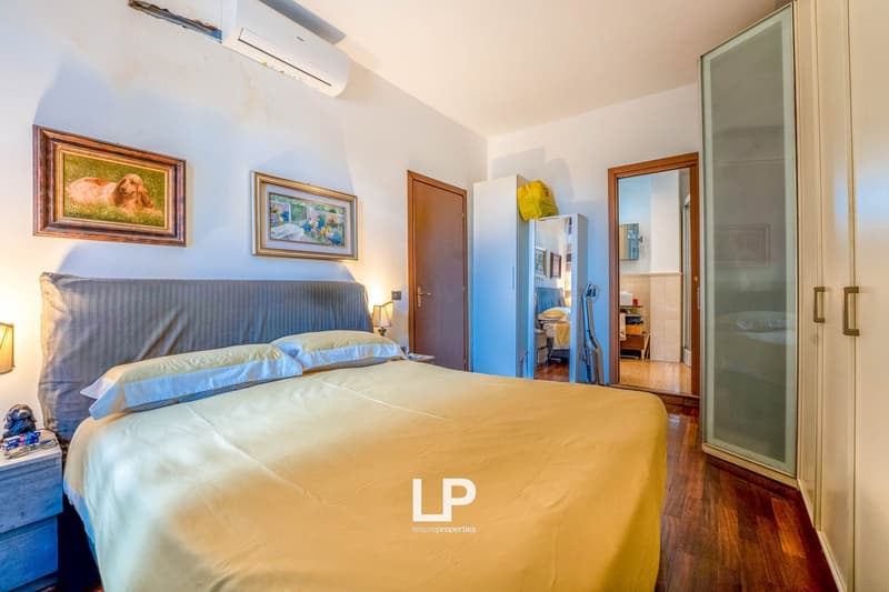 Spacious bedroom with hardwood floors, white walls, large bed with blue and yellow bedding, framed artwork on the walls, air conditioning unit, and a private washing machine visible in the room.