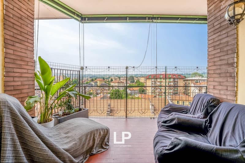 Spacious balcony with glass railing, overlooking a city skyline. The balcony features potted plants, a couch, and a view of the surrounding buildings and landscape.