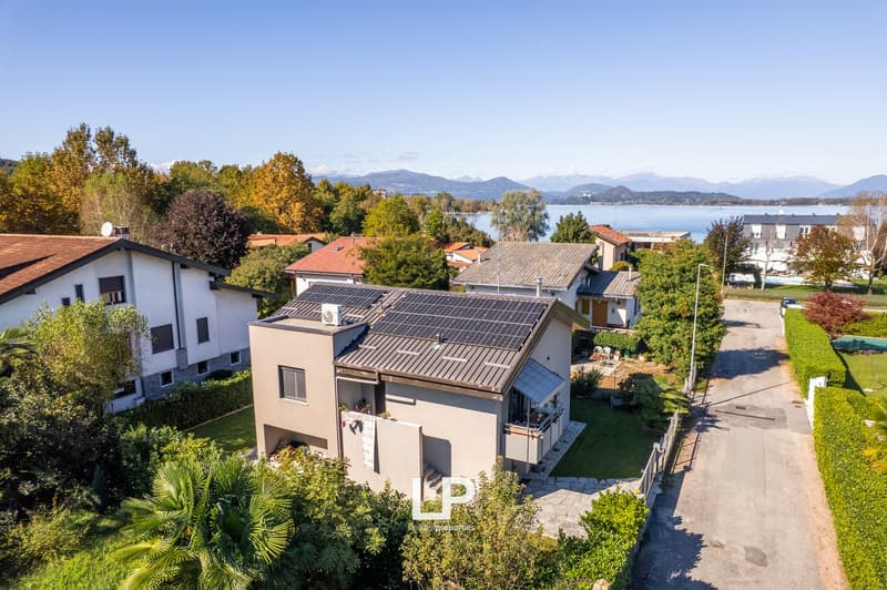 Detached house with solar panels on the roof, surrounded by trees and grass, with a driveway leading to it, mountains and a lake in the background.