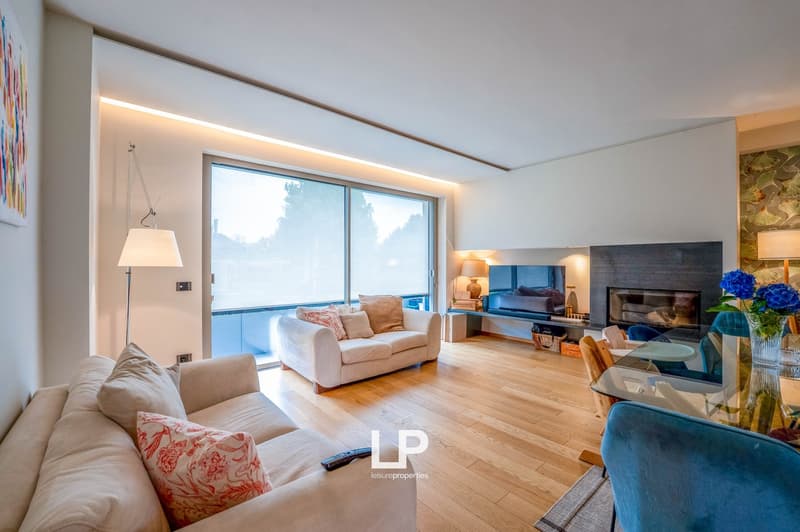 Spacious living room with large windows providing a scenic view, hardwood flooring, white sofas, fireplace, and decorative accents such as artwork and floral arrangements.