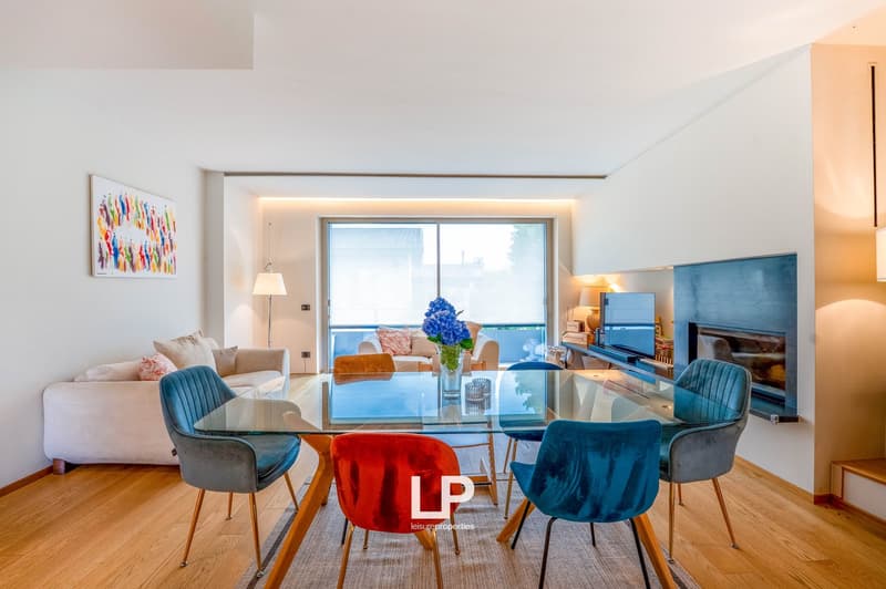 Spacious living room with large windows, modern furniture including a glass dining table, colorful chairs, and a sofa. There is a balcony or terrace visible through the windows.