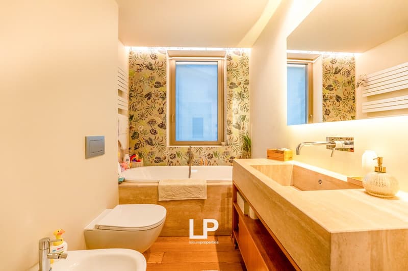 Spacious bathroom with floral wallpaper, wooden vanity with sink, toilet, and large window providing natural light
