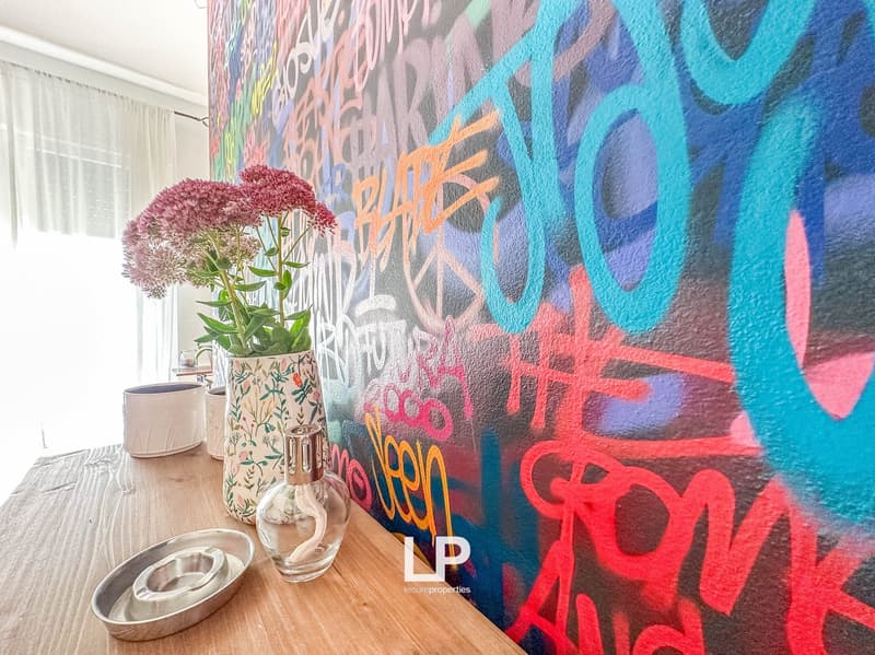 The image shows a room with a colorful graffiti-style mural on the wall, featuring vibrant colors and abstract patterns. There are vases with flowers on a wooden table, and a window with curtains in the background, suggesting the presence of a balcony or