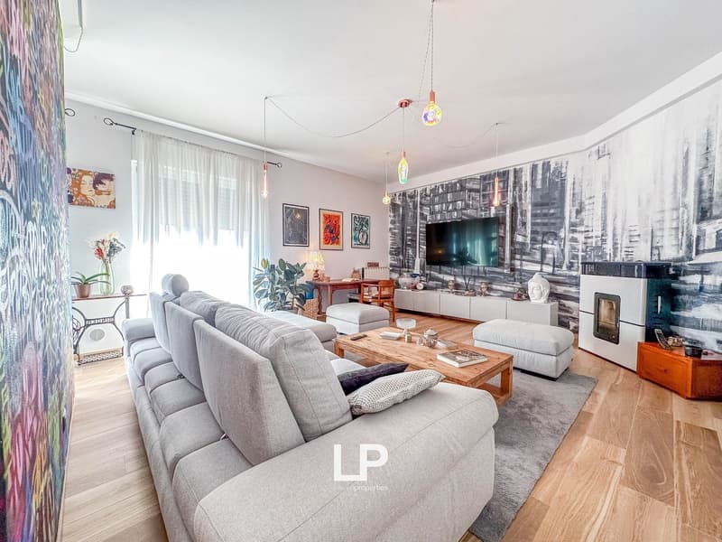 Spacious living room with gray sofas, wooden coffee table, wall-mounted TV, colorful artwork, and a fireplace. The room has a modern and eclectic decor with pendant lights and potted plants.