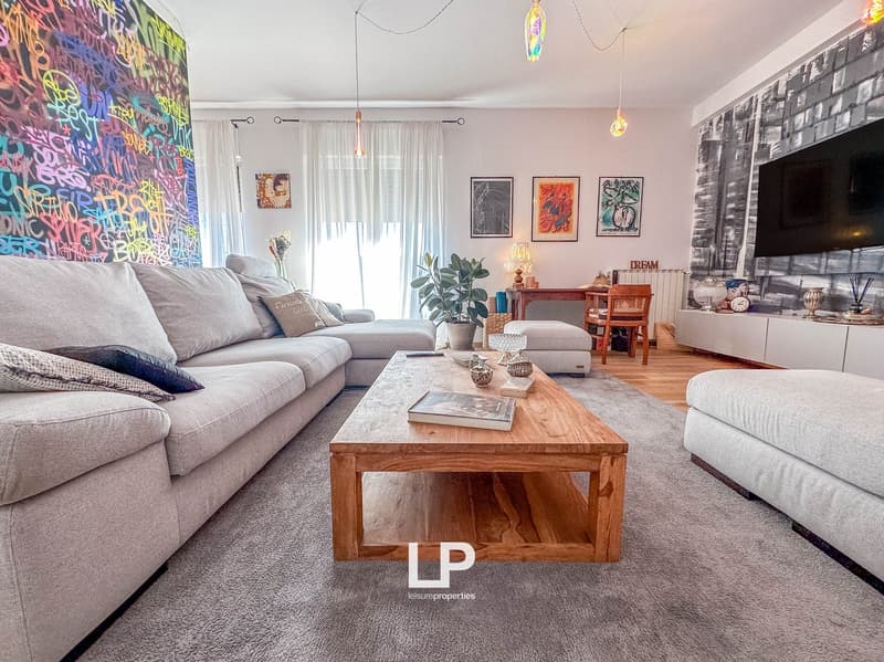 Spacious living room with gray sectional sofa, wooden coffee table, wall decor, potted plant, and hanging light fixtures. The room features a colorful graffiti-style mural on one wall and a fireplace.