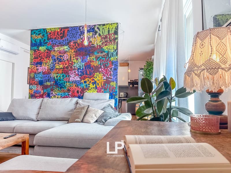 Colorful graffiti-style wall art, gray sectional sofa, wooden coffee table, potted plants, macrame wall hanging, and a fireplace in a modern, eclectic living room setting.