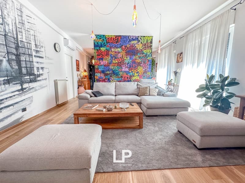 Spacious living room with large gray sectional sofa, wooden coffee table, and colorful graffiti-style wall art. The room also features a large window, hardwood floors, and a potted plant.