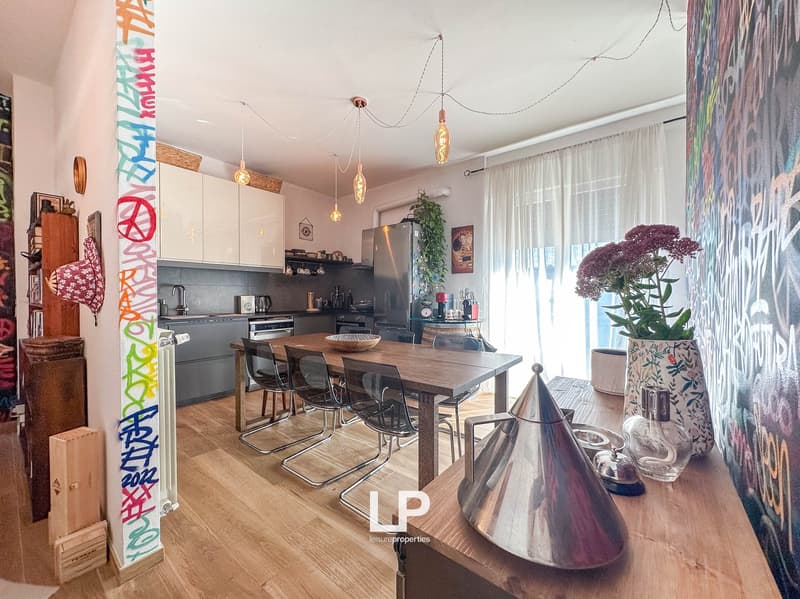 Open kitchen with wooden dining table and chairs, pendant lighting, colorful wall decor, potted plant, and large windows