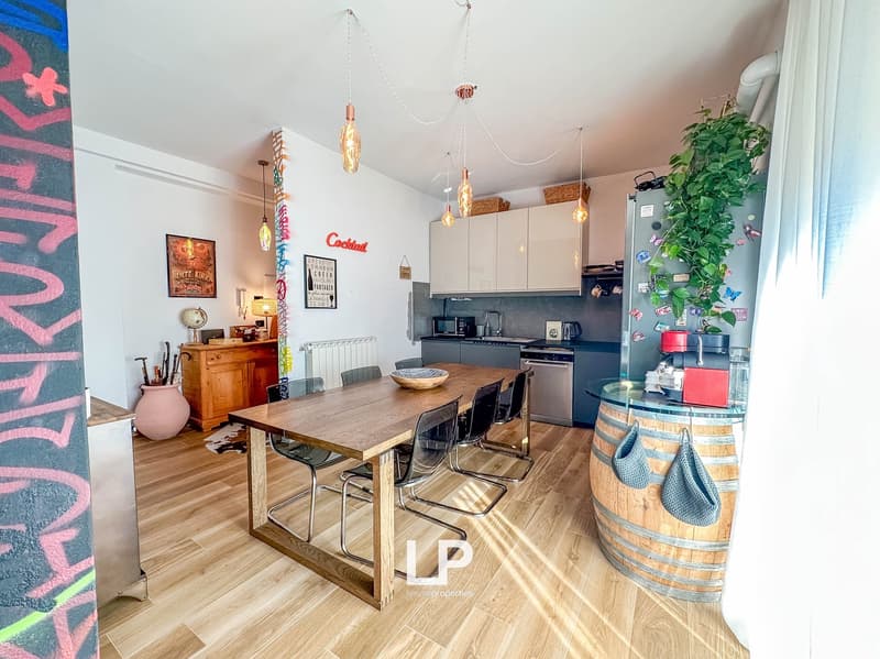 Open-concept kitchen and dining area with wooden dining table and chairs, kitchen cabinets and appliances, hanging pendant lights, and decorative elements like a barrel and potted plant